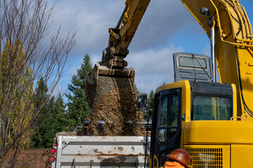 Excavator bucket attachment spilling dirt into a dump truck to haul away, public infrastructure construction site
