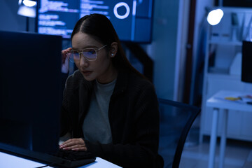 Serious Asian female programmer coding software developer focused on laptop screen working late night deadline with blue light showing stress and concentration in dark modern office environment