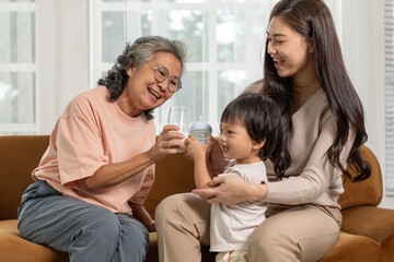 Three generations of happy asian family relaxing on brown sofa at home featuring smiling grandmother giving water to cute grandson held by cheerful mother bonding together with love care and joy