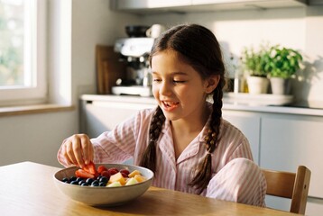 Happy girl with braids eating fresh fruit salad bowl in sunny kitchen wearing pink striped pajamas