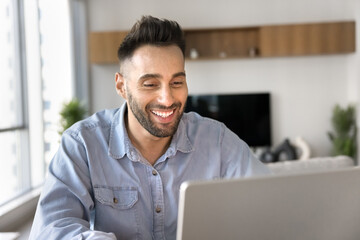 Smiling man using laptop seated at table at home office, engaged in online meeting or webinar,...