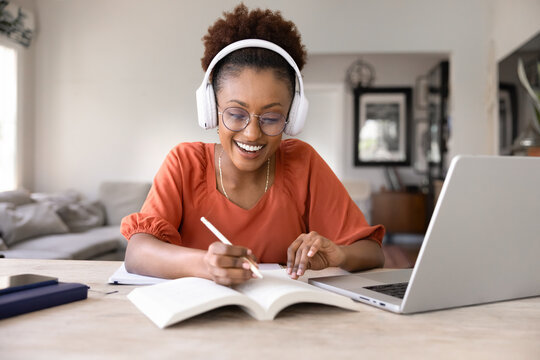 Cheerful young Black student woman in eyeglasses and headphones listening to learning webinar at laptop, writing notes in copybook, doing research study, enjoying online education at home