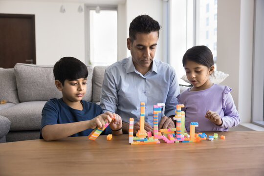 Mature 4s Indian father spending quality time with his little son and daughter while building creative structures using colorful toy bricks at home. Educational play, bonding, imagination development