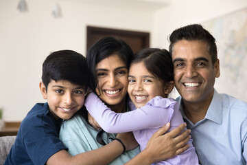 Close up portrait of loving Indian parents with cute little kids hugging seated on sofa at home,...