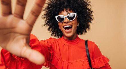 A smiling woman with curly hair in a red top and white sunglasses joyfully reaches towards the camera against a tan backdrop. She exudes confidence and a positive attitude.