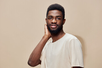 Young African man in a light t shirt, smiling and touching his neck, with a neutral beige...