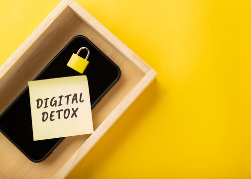 High angle of a smartphone in a wooden tray with a yellow padlock and a sticky note saying Digital Detox. Concept for social media addiction, mental health, and technology disconnect for wellbeing.
