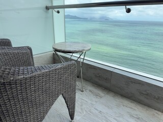 Obraz premium Ocean View Balcony: A patio chair and table on a glass balcony overlooking beautiful blue-green ocean waters.