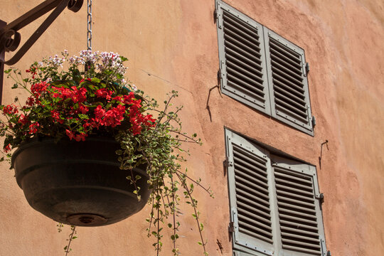 Close-up of a hanging flower pot on a warm-toned Mediterranean wall with traditional window shutters, ideal for lifestyle, home, and travel concepts.