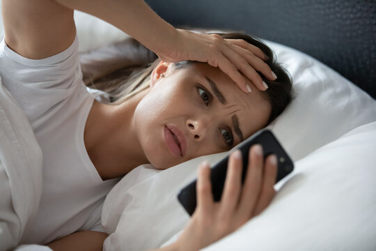 Morning phone stress. Distressed nervous millennial woman lying in bed checking smartphone after waking up hug head looking at screen missed important call read stressful text message overslept alarm