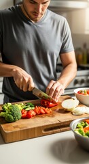 Man in a gray t-shirt chopping vegetables on a wooden cutting board in a kitchen with a bowl of salad.