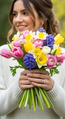 Woman holding a colorful bouquet of flowers with pink, yellow, white, and purple blooms in her hands, wearing a white sweater with a blurred green background.