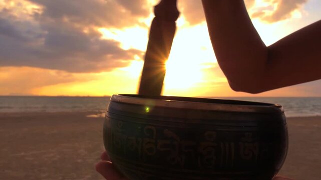 Silhouette of a person playing a traditional tibetan singing bowl with a mallet on a tranquil beach, embracing a spiritual moment as the sun sets over the serene ocean horizon