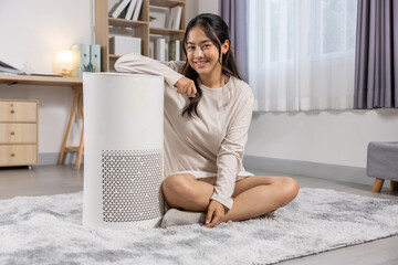 Happy young asian woman smiling sitting on rug with white air purifier enjoy fresh clean air in...
