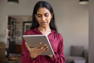 Positive young Indian gadget user woman typing on tablet at home, browsing Internet, social media, checking email, managing remote work business process, standing and holding digital device