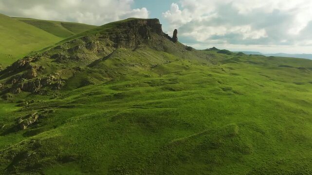 An aerial drone shot captures the rugged, green landscape of the Trotternish peninsula on the Isle of Skye, Scotland, featuring the iconic Old Man of Storr rock formation under a cloudy sky.