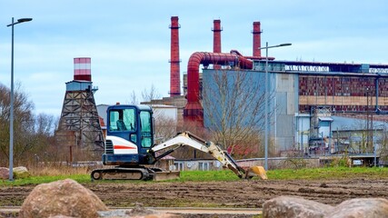 Small excavator working on a construction site near an industrial plant with factory buildings and smokestacks. Concept of urban development, industry, infrastructure, land preparation, renovation, 