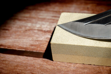 Knife blade resting on a whetstone block, getting sharpened on a wooden background. Maintenance tool for sharp edge