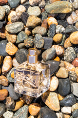 Close-up of a transparent square perfume bottle against the background of the pebbles of the beach. A blank bottle mockup. Presentation sea aroma