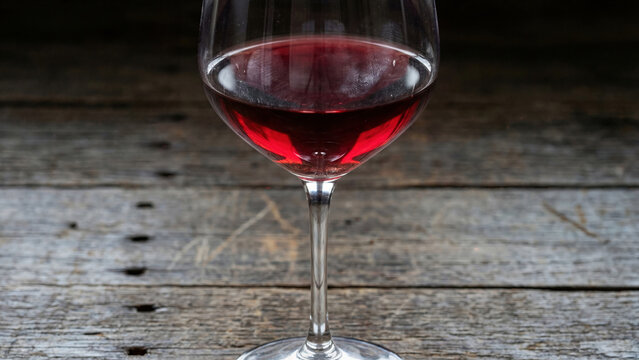 Artistic macro photograph of deep red wine in elegant Bordeaux glass on weathered barn wood table, wine catching light creating ruby red translucency, finger marks visible on glass bowl showing human