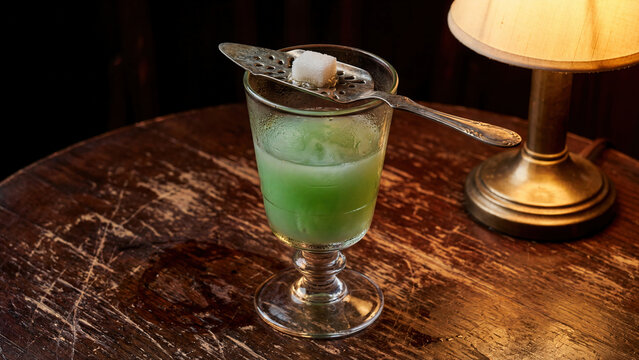 Artistic macro shot of absinthe in traditional Pontarlier reservoir glass with decorative etched dose line, liquid showing characteristic green opalescent louche effect from water addition, antique
