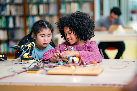 Black girl collaborates with Asian girl during robotics learning session, connecting wires, assembling robot parts, STEM education focus, diversity, teamwork skills, technology curiosity in classroom. - Powered by Adobe