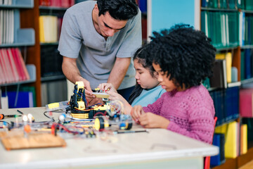 Teacher assists children during robotics build session, sharing technical knowledge, electronics practice, STEM learning experience, cooperative classroom interaction, guided education moment.