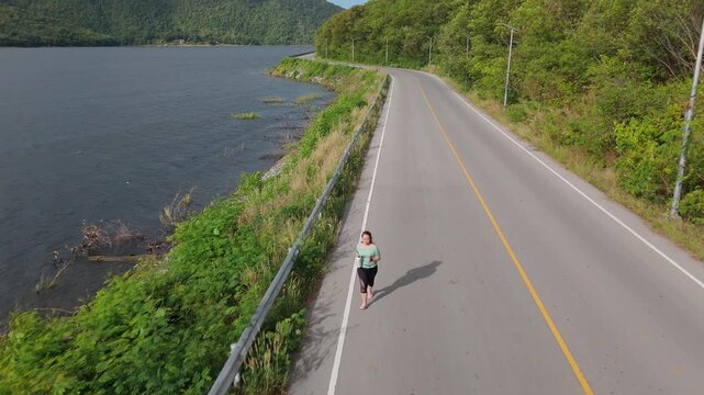 Asian woman jogging on a quiet road beside a lake, holding a reusable water bottle in a natural outdoor environment. Sustainable lifestyle, and a healthy fitness routine.