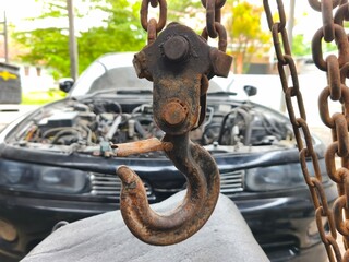 Close-up of a Heavy-Duty Rusty Industrial Hoist Hook and Chain with a Blurred Car Engine Background, Representing Automotive Repair, Vehicle Towing Service, and Workshop Engineering