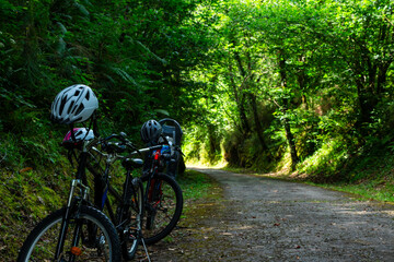 Two bikes are parked on a dirt road in a forest. The bikes are both black and have helmets on them. The helmets are placed on the handlebars of the bikes. The bikes are parked next to each other