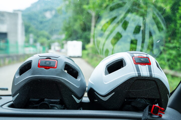Two helmets are on the dashboard of a car. One is black and the other is white. The helmets are placed side by side, and the car is driving down a road
