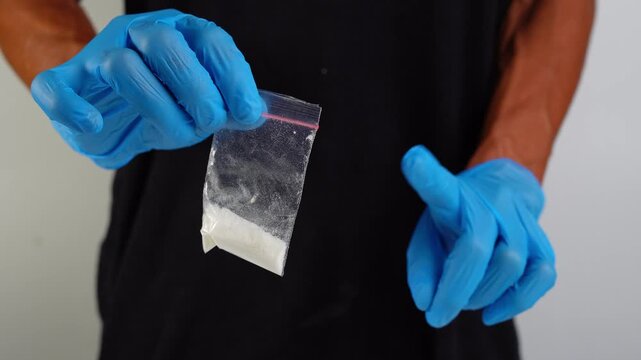 A man wearing blue protective gloves holding small plastic bag containing white powder substance, illustrating illicit drug evidence or handling