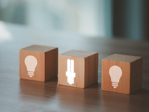 Wooden cube with glowing energy saving light bulb icon next to two wooden cubes with light bulb icons representing sustainable energy choice and innovation in modern workspace