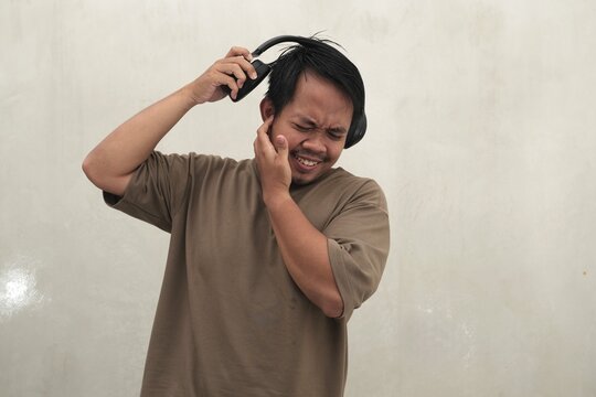 Southeast Asian man wearing casual t-shirt and headphones reacting with earache and discomfort, highlighting hearing problems, loud sound issues, and poor audio experience for health