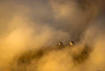 Two small houses sit on a hill as clouds cover them in the sunset light. The landscape has a soft glow, creating a quiet scene of nature.