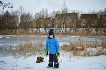 Child boy standing  in winter coat with snowy background near river in residential area.