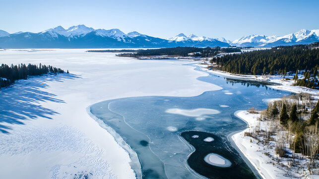 A vast frozen lake lies amidst a snow-covered forest, overlooking a mountain range on a clear winter day - Powered by Adobe