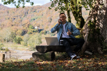 Fototapeta premium Young man gives thumbs up while on call with coffee and laptop in fall nature
