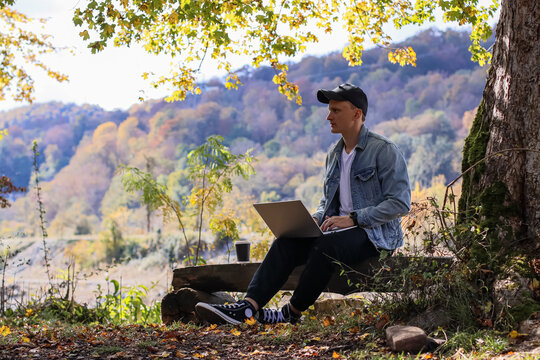 Man in cap works on computer outdoors in colorful autumn woods with river in distance