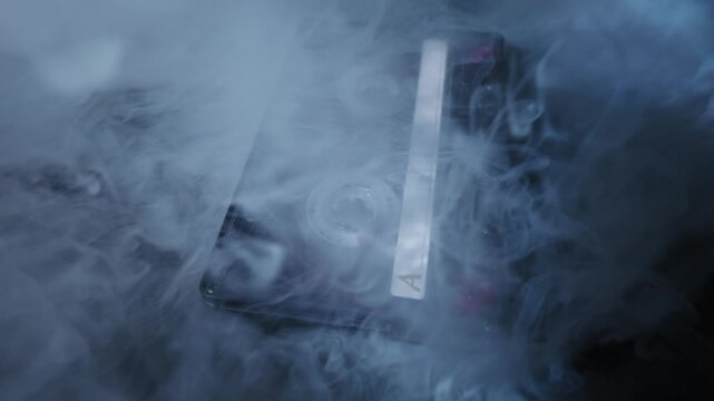 Transparent audio cassette tape with Side A label lies on a dark surface. Thick, white, atmospheric smoke billows across the frame, creating a mysterious mood.