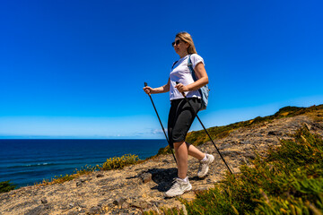 Beautiful middle-aged woman tourist hiking on trail along Atlantic ocean in nature park Vicentine Coast in Algarve in Portugal on summer day. Side view