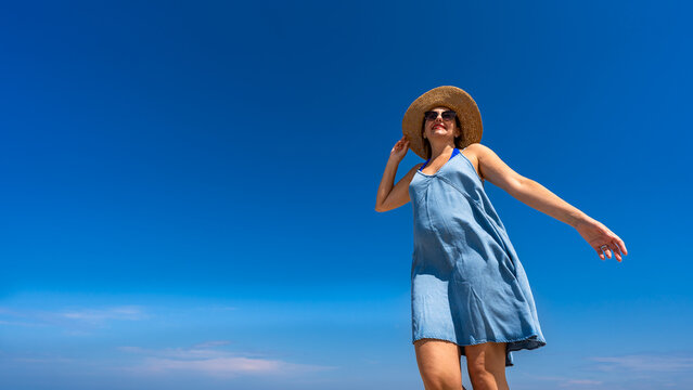 Portrait of beautiful happy middle-aged woman in summer dress and sun hat standing against background of blue sky in summertime. Front view