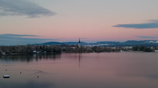 Aerial drone view of Cham on Lake Zug in winter, where a snow-covered landscape meets calm water at dusk, creating a quiet and atmospheric evening scene.