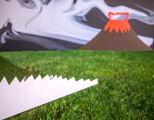 Cardboard and paper volcano erupts over grassy surface, abstract background