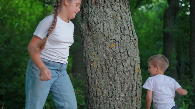 Boy chases girl around tree playing tag in forest outdoors. Children run circling trunk during game. Kids play chase in forest. Boy runs after girl around tree. Children enjoy tag game in nature.