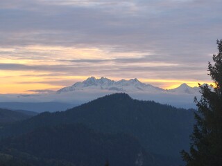 This breathtaking landscape captures the majestic beauty of snow-capped mountain peaks during a vibrant sunrise. The composition features a deep blue foreground of forested hills, transitioning into a
