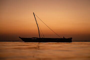 A traditional wooden dhow sailing boat is silhouetted against a calm sea and a deep orange sky during sunset in Zanzibar.