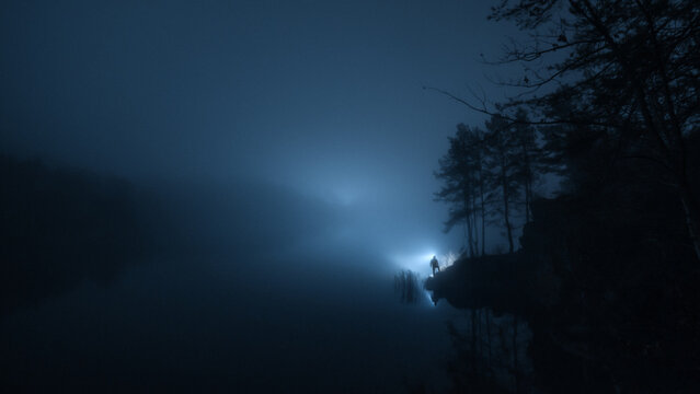 Silhouette of a lone person with flashlight standing by foggy lake at night. Mysterious dark landscape, loneliness and journey concept, fear of unknown, dramatic atmosphere, copy space.