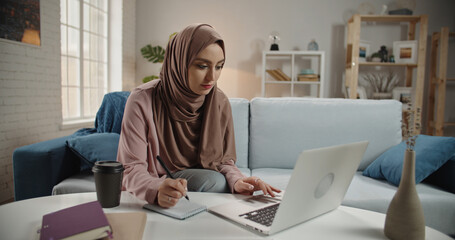 Focused Muslim Woman in Hijab Writing Notes While Looking at Laptop Screen Online Learning Remote Work and Education Concept