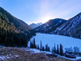 Setting sun in the mountains. Frozen lake. Wild forest on the slopes. Drone view.